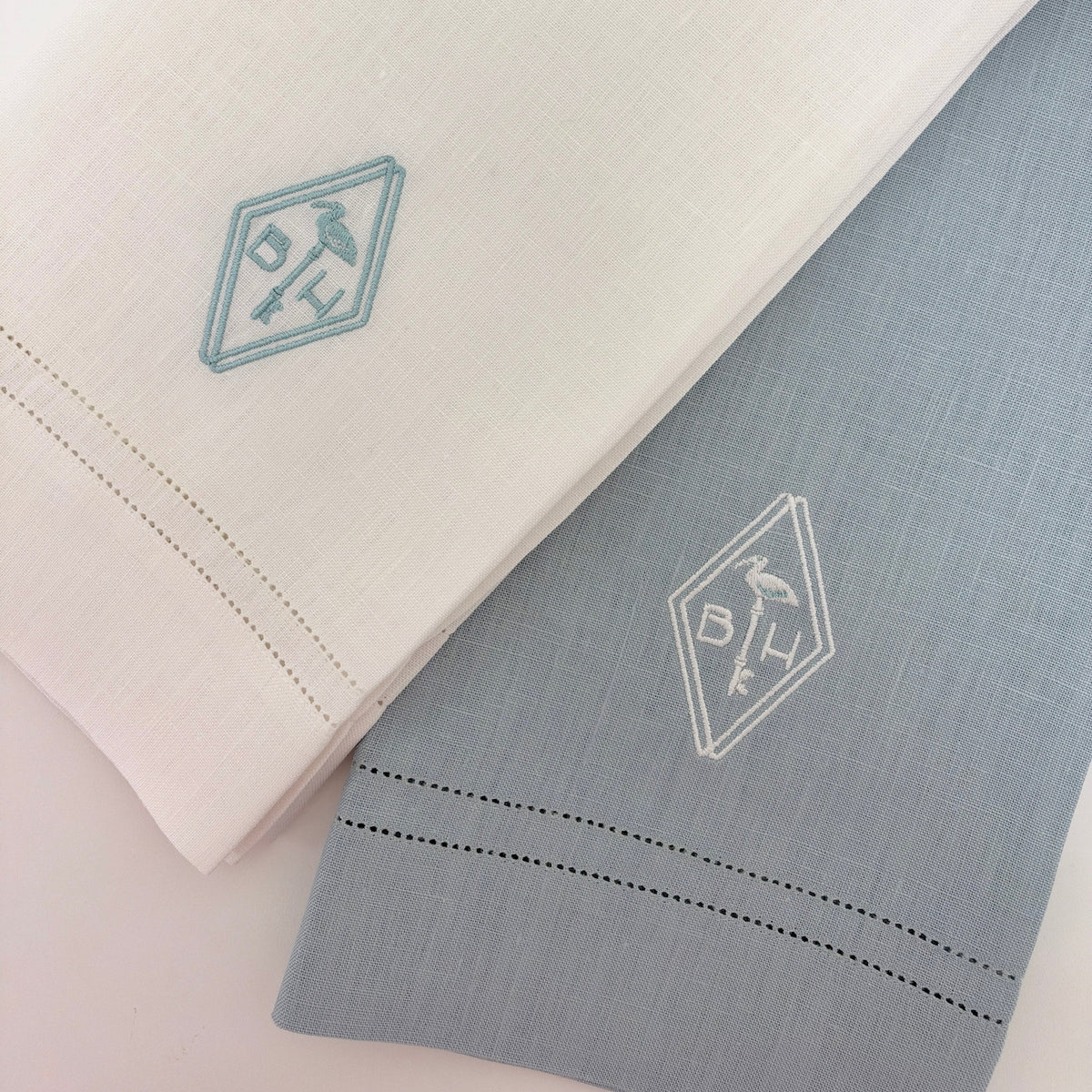 Two folded embroidered cloth napkins or handkerchiefs on a white background, one in white with a light blue embroidered logo and hemstitch, and one in pale blue with a white embroidered logo and hemstitch.
