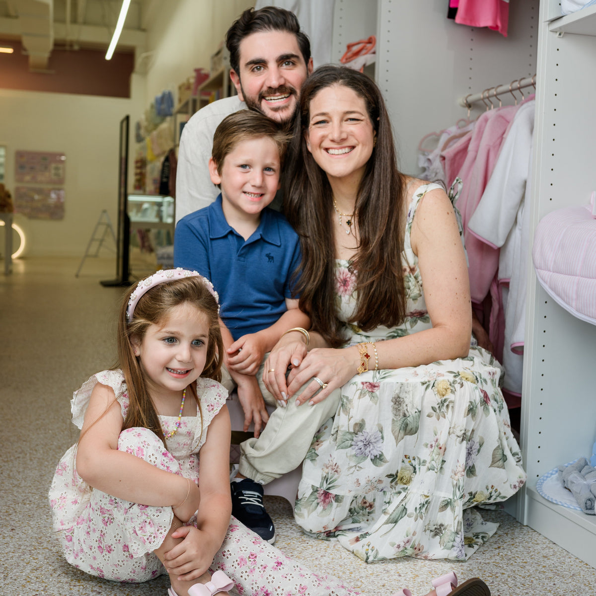 A family of four posing together in a retail store clothing section. The mother wears a sleeveless floral print maxi dress, the father wears a light-colored polo shirt, the young boy wears a blue polo shirt, and the young girl wears a matching floral romper and sandals.